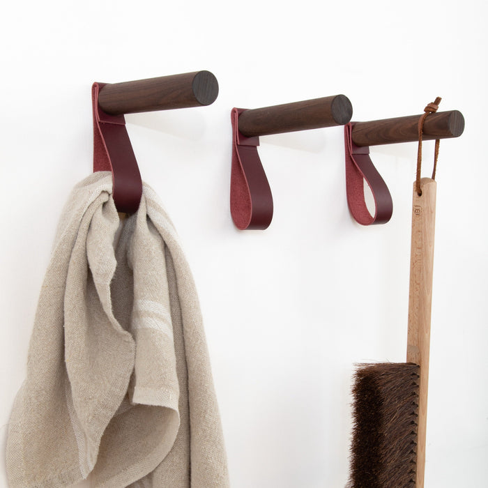 Keyaiira Wine colored leather wall strap on dark walnut wooden peg holding a neutral linen towel, handcrafted minimalist wall hook for bathroom, laundry room, or entryway organization.
