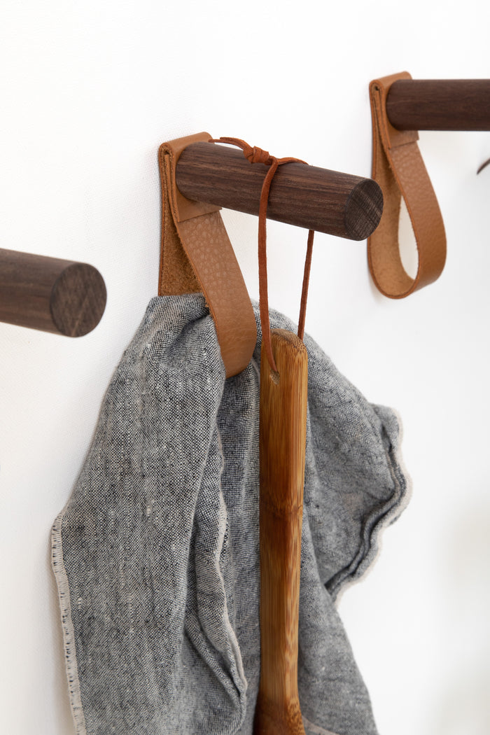 Keyaiira nicotine leather wall strap mounted on a dark walnut peg holding a gray linen cloth and natural bristle cleaning brush, handcrafted minimalist wall hook for modern kitchen or utility room organization.