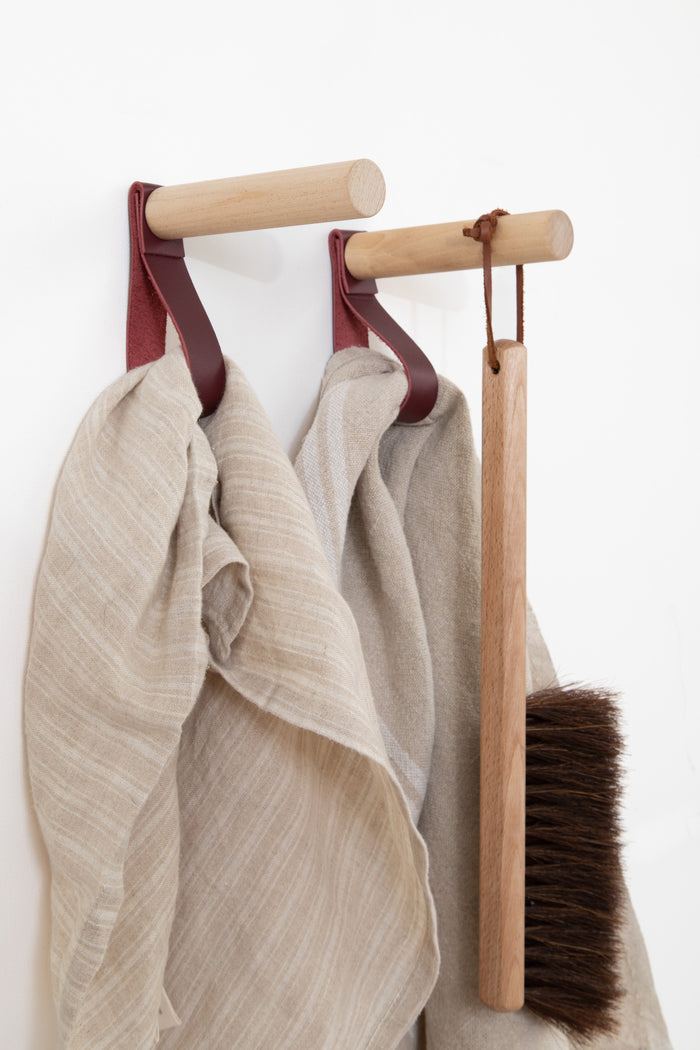 Wine-colored leather wall peg hooks holding linen towels and a wooden cleaning brush — handcrafted minimalist home organizer with natural wood pegs and vegetable-tanned leather straps.