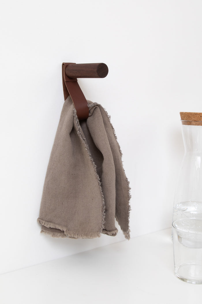 Walnut wood wall peg with walnut brown leather strap holding a neutral linen towel handcrafted modern wall hook for minimalist home organization.